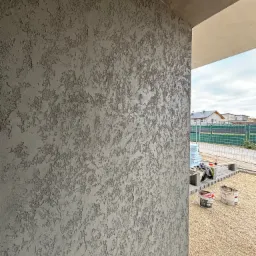 Szara ściana zewnętrzna z efektem dekoracyjnym baranka, widoczne wiaderka farby i przygotowane materiały budowlane w tle.