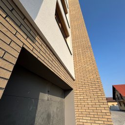 WellDone W&B Bartosz Wójcik - Nowoczesna elewacja budynku z jasnej cegły klinkierowej i białego tynku, widok z dołu na fragment ściany z bramą garażową w kolorze antracytowym.