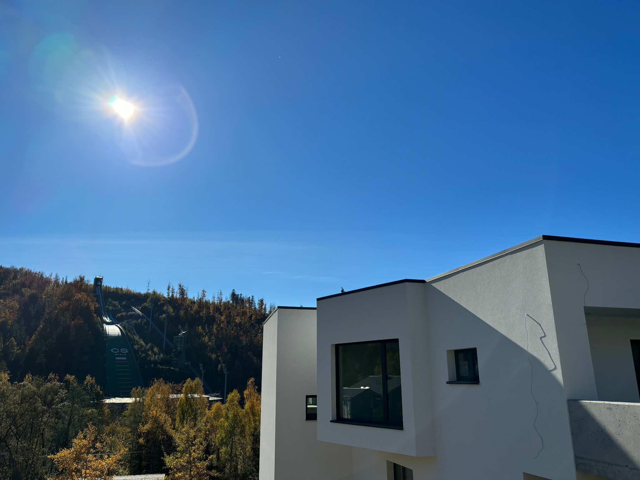 Nowoczesny, biały budynek z płaskim dachem na tle zalesionych wzgórz i skoczni narciarskiej w słoneczny dzień. Widoczne minimalne pęknięcia na elewacji.