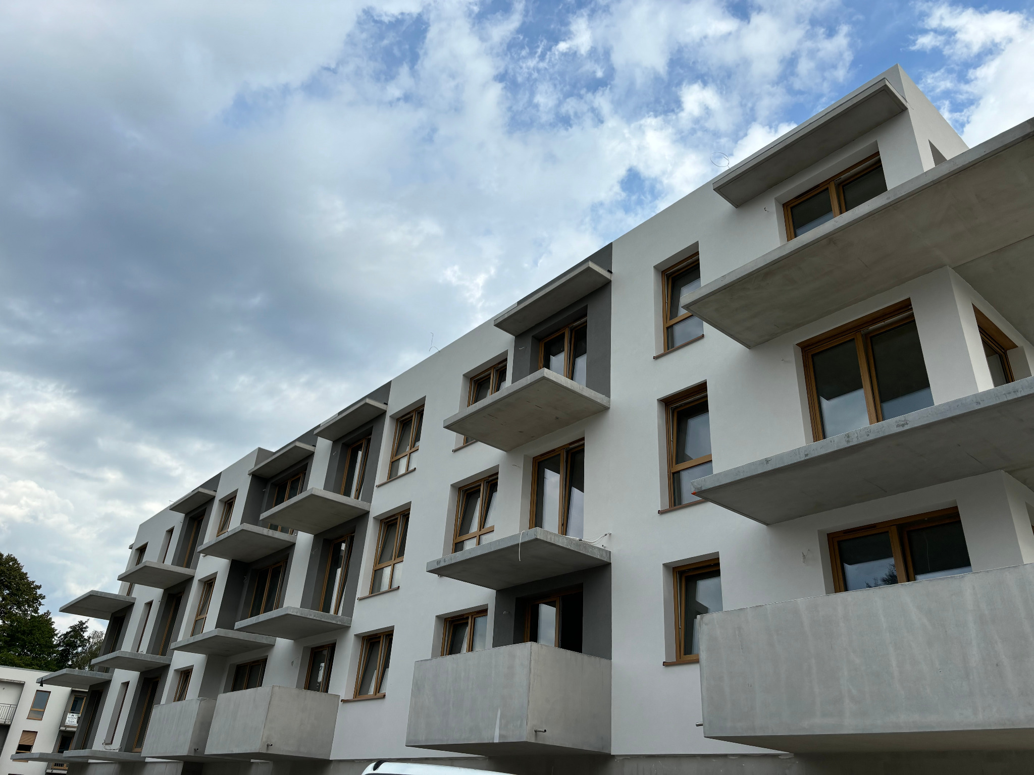 Nowoczesny budynek wielorodzinny z białymi ścianami i szarymi akcentami, widoczne balkony z surowego betonu i drewniane ramy okienne, pochmurne niebo w tle.