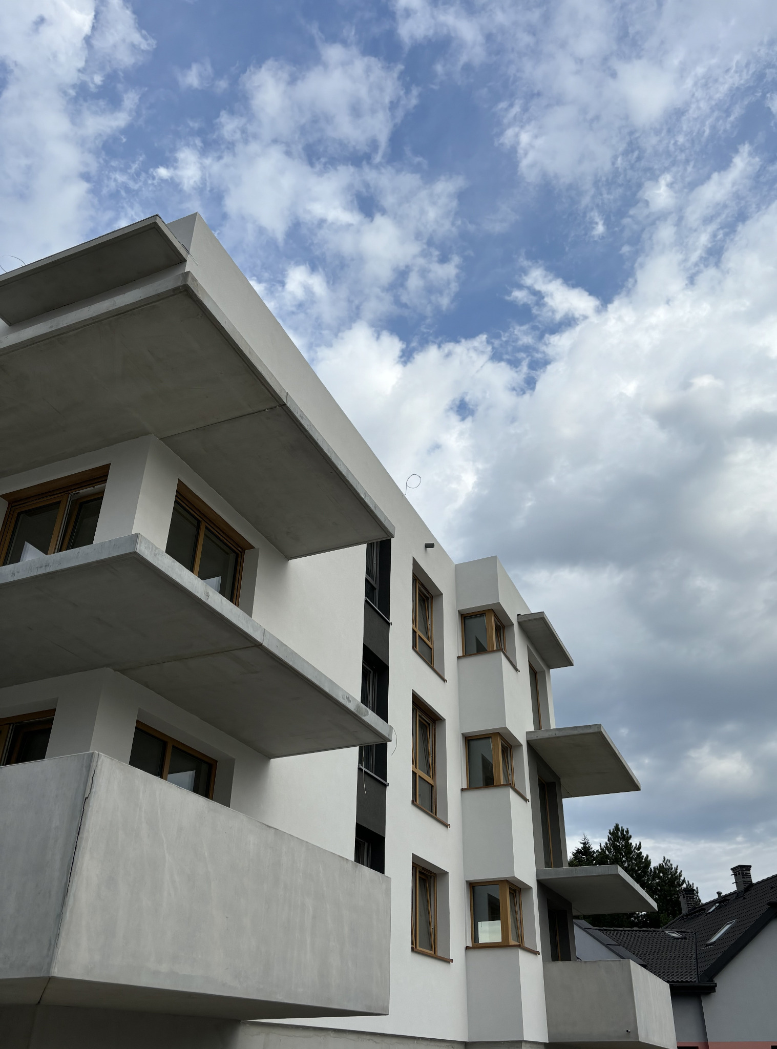 Nowoczesny budynek wielorodzinny z białymi ścianami i betonowymi balkonami, sfotografowany pod kątem w pochmurny dzień.