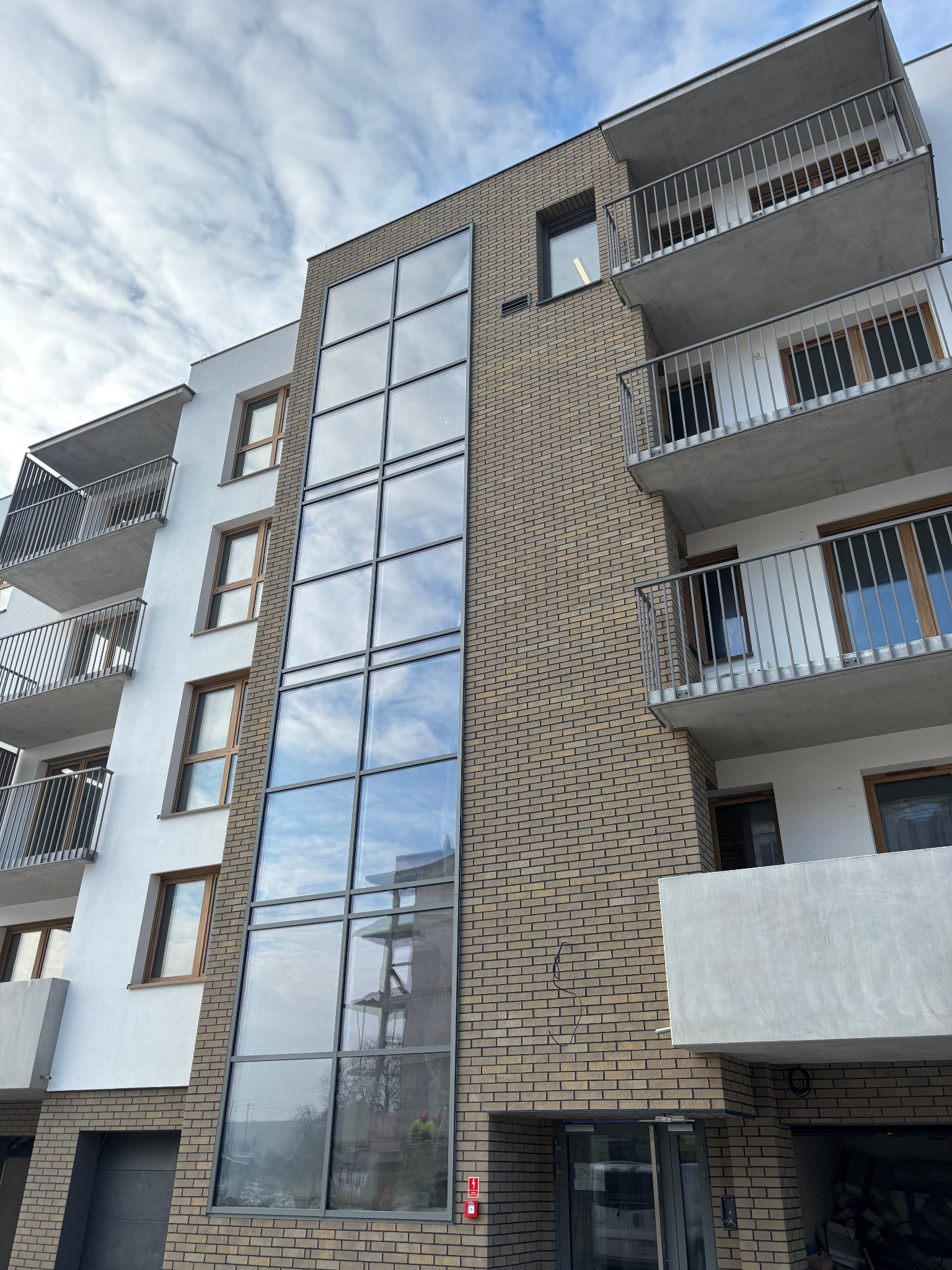 Nowoczesny budynek wielorodzinny z elewacją z cegły klinkierowej i dużymi oknami odbijającymi chmury, widok z dołu na fragment fasady z balkonami.