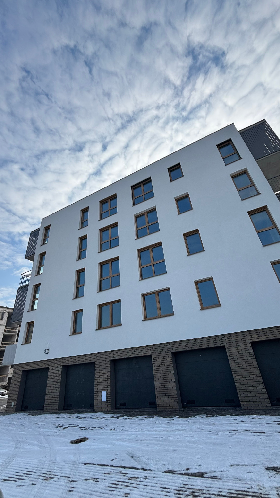 Nowoczesny budynek wielorodzinny z białą elewacją i brązowymi oknami, widok z dołu na tle pochmurnego nieba, z garażami na poziomie gruntu wykończonymi cegłą klinkierową i śniegiem na pierwszym...