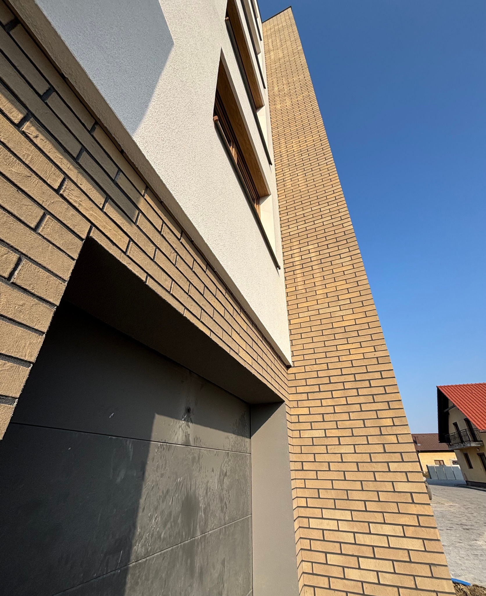 Nowoczesna elewacja budynku z jasnej cegły klinkierowej i białego tynku, widok z dołu na fragment ściany z bramą garażową w kolorze antracytowym.
