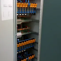 Otwarta szafa elektryczna z widocznymi bezpiecznikami, przewodami i listwami zaciskowymi, etykiety na urządzeniach, schemat na drzwiach.