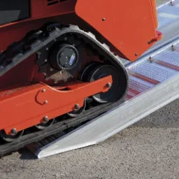 Pomarańczowa miniładowarka gąsienicowa wjeżdża na aluminiową rampę, widok z boku na gąsienice i rampę.