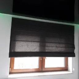 Ciemna, poziomo składana przesłona okienna w drewnianej ramie, z widocznym systemem sterowania sznurkowym, w nowoczesnym wnętrzu z oświetleniem LED w narożnikach sufitu.