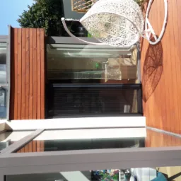 Zamknięta, czarna, pozioma przesłona okienna w nowoczesnym domu z drewnianą elewacją i wiszącym fotelem ogrodowym, rzucającym ażurowy cień na taras.