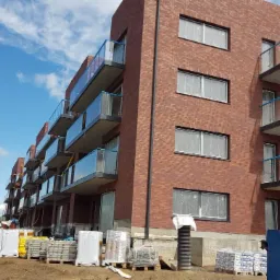 Nowo wybudowany budynek z czerwonej cegły z balkonami z szklanymi balustradami i czarnymi ramami okiennymi, okna zabezpieczone folią, widoczne materiały budowlane na placu budowy.