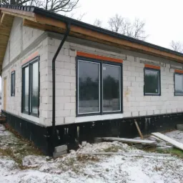 Surowy budynek w trakcie budowy z zamontowanymi oknami z czarnymi ramami, widoczne bloczki białe i czarna izolacja fundamentu, zimowa sceneria z resztkami śniegu na ziemi.