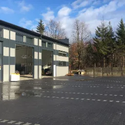 Zewnętrzny widok magazynu z otwartymi bramami, widoczne regały i sprzęt wewnątrz, duży parking przed budynkiem, w tle las.