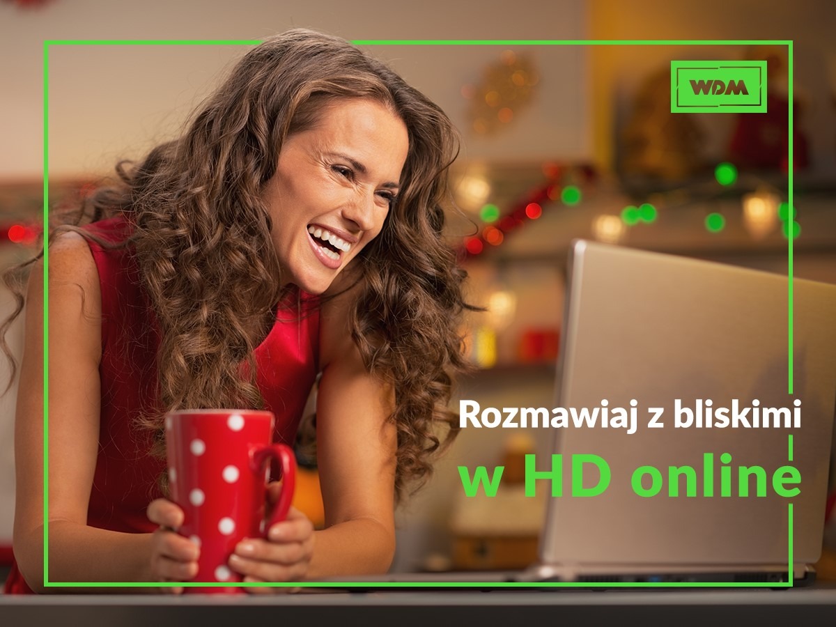 Uśmiechnięta kobieta z kręconymi włosami w Mielcu, w czerwonej sukience, trzyma czerwony kubek w białe kropki, rozmawiając przez wideokonferencję na laptopie; w tle dekoracje świąteczne i logo firmy.