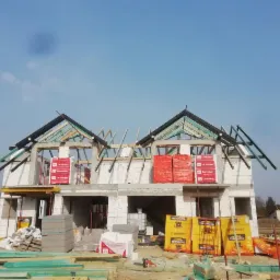 Wznoszony dwukondygnacyjny dom z bloczków białych, z widoczną konstrukcją dachu i przygotowanymi materiałami izolacyjnymi Termobet na balkonach, oraz paletami z bloczkami Termalica na placu budowy.