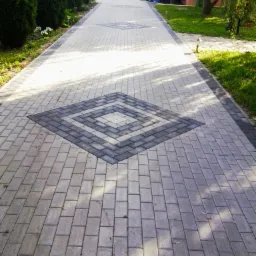 Długa, prosta ścieżka z jasnoszarej kostki brukowej z ciemnoszarymi wstawkami w kształcie rombów, prowadząca do domu z wózkiem na posesji w tle.