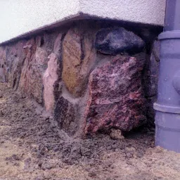 Kamienna podmur&oacute;wka budynku z widoczną rurą spustową, ziemia wok&oacute;ł fundament&oacute;w.