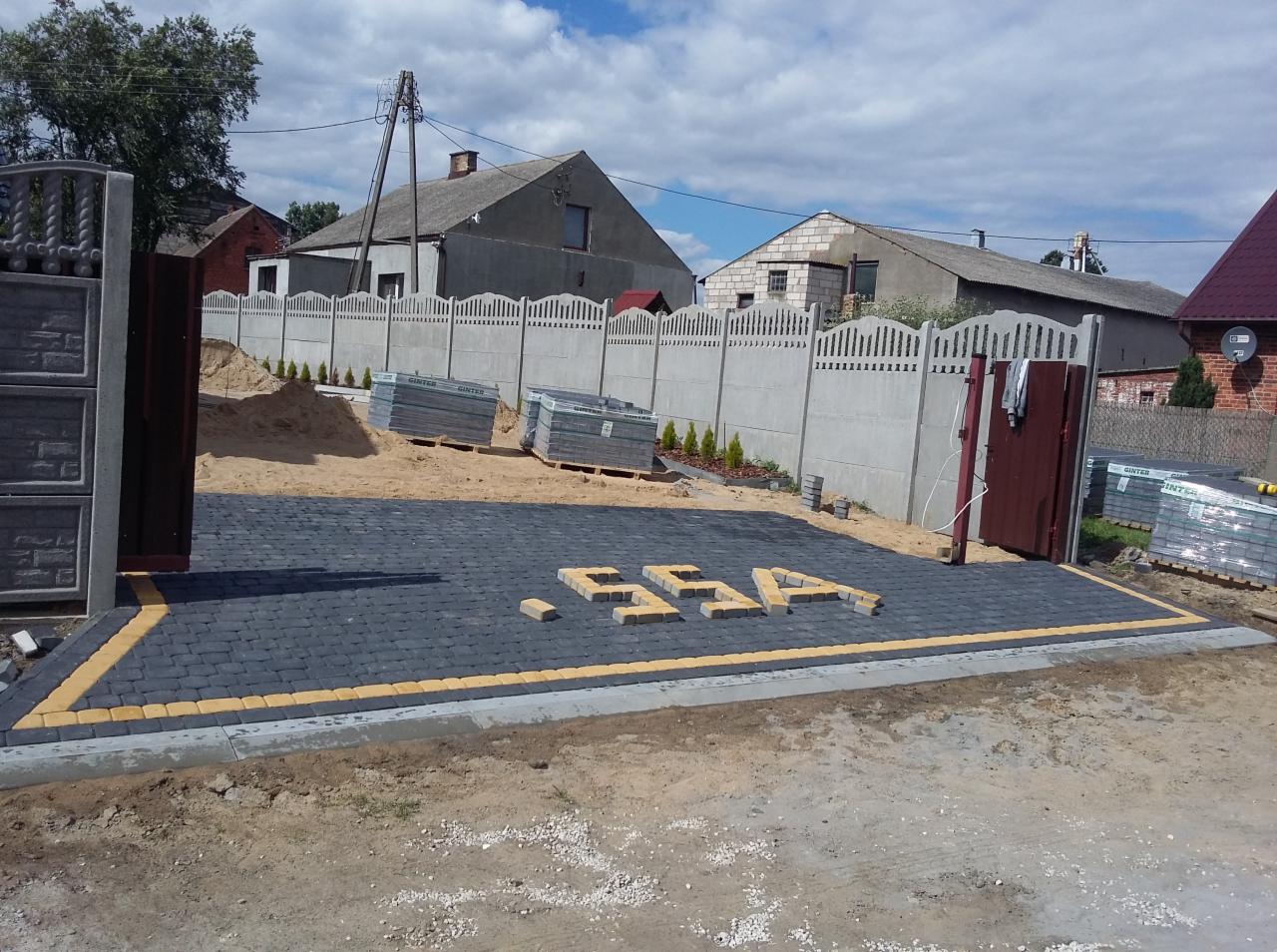 Częściowo ułożona, ciemnoszara kostka brukowa z żółtymi obrzeżami, z ułożonym napisem 'SSA' z żółtej kostki, na tle betonowego ogrodzenia, palet z kostką i budynków.