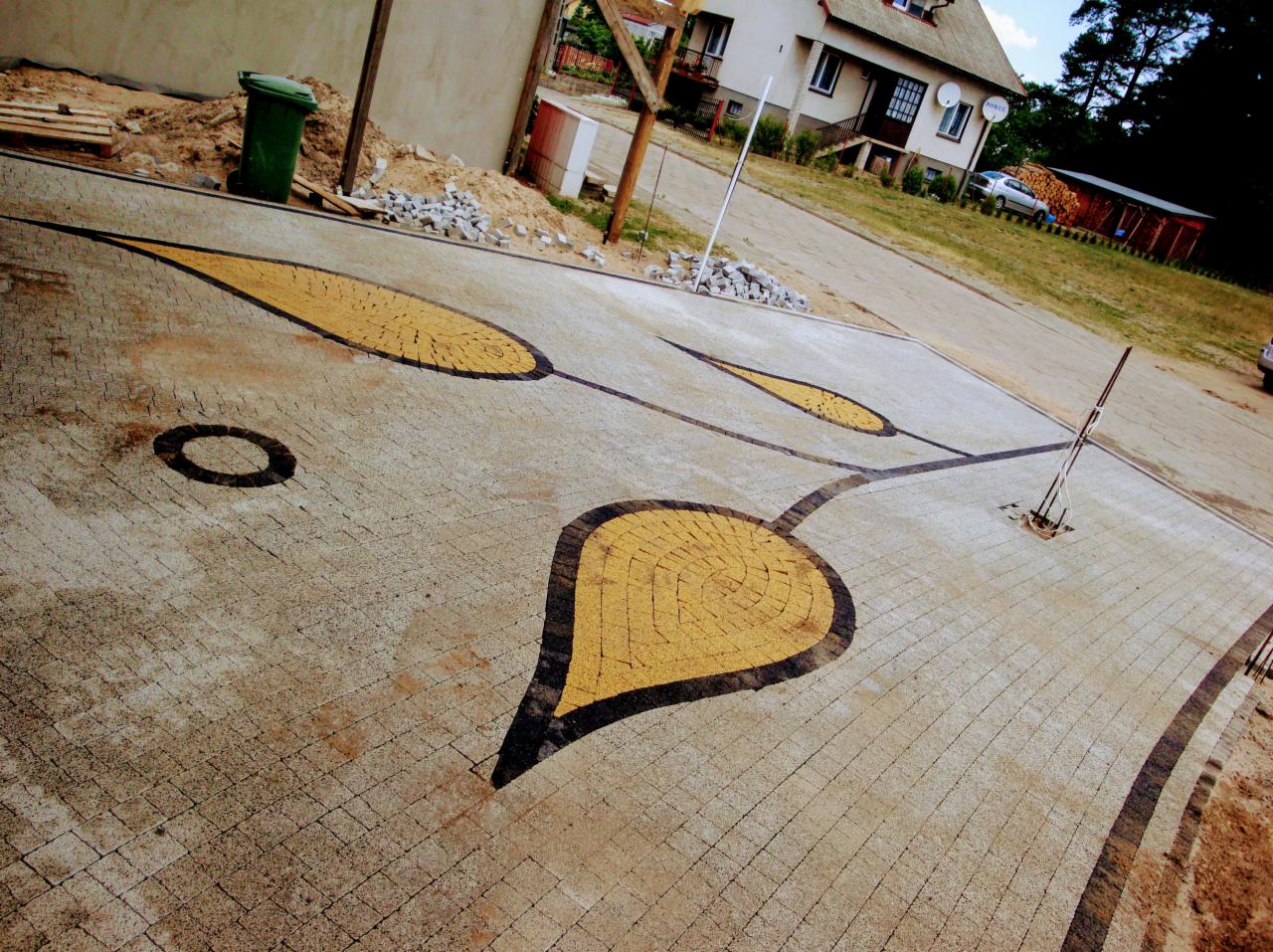 Ukończona nawierzchnia z kostki brukowej z dekoracyjnym wzorem liścia w kolorze żółtym otoczonym czarną obwódką na szarym tle, widoczny budynek w tle.