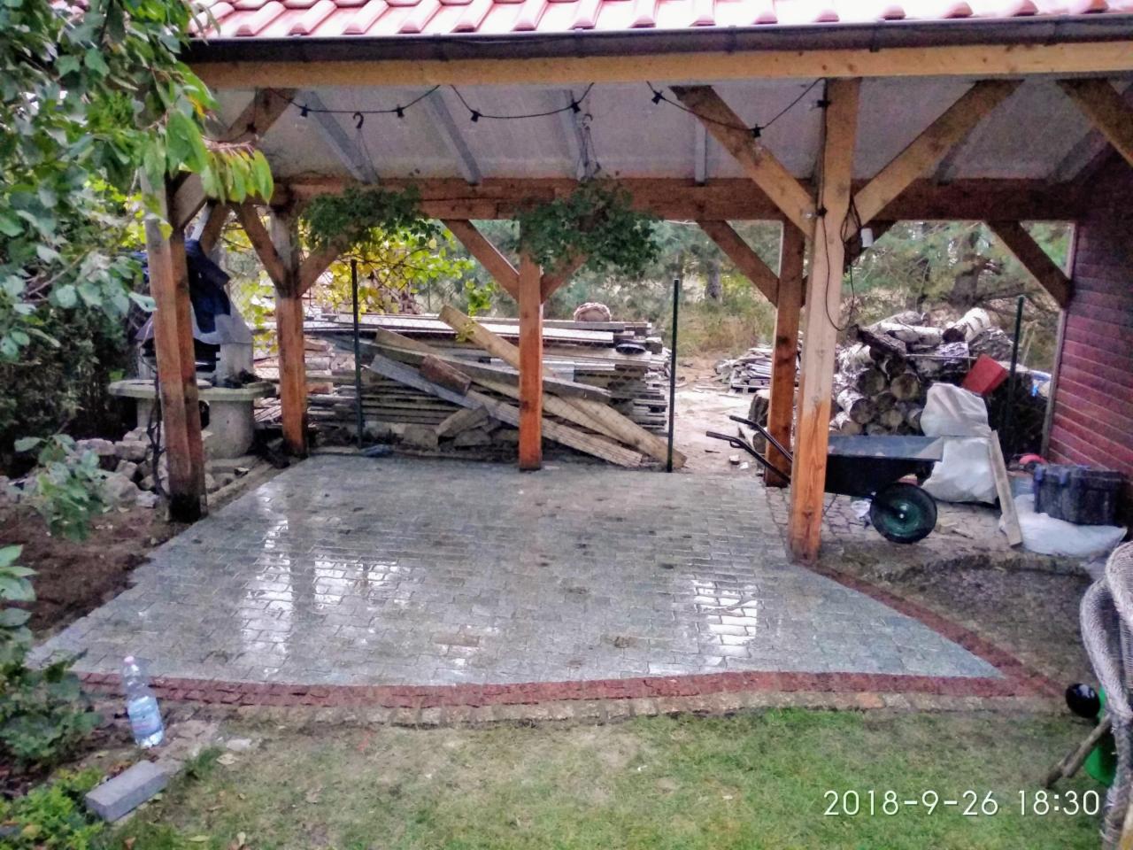 Świeżo ułożona, mokra kostka brukowa w altanie z drewnianą konstrukcją, obrzeże z czerwonej kostki, w tle składowane drewno i narzędzia.