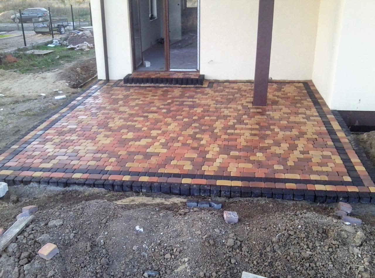 Ułożona kostka brukowa w odcieniach czerwieni, pomarańczy i żółci z czarnym obramowaniem na patio przy domu, widoczne niedokończone prace ziemne.