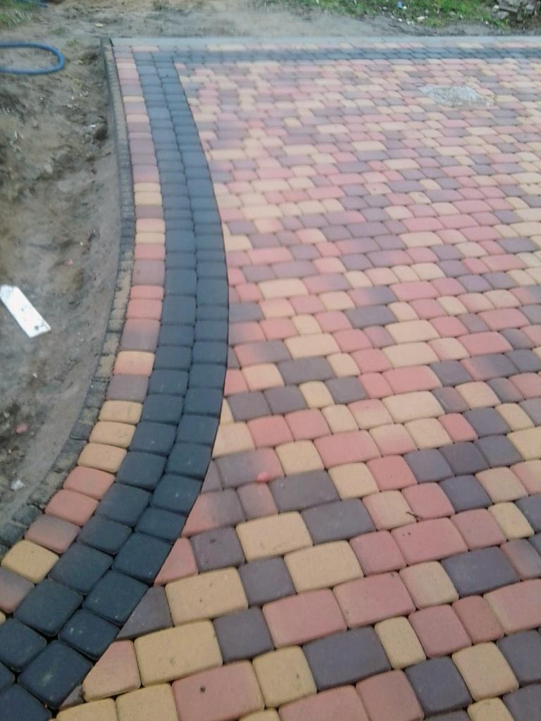 Ułożona nawierzchnia z kostki brukowej w odcieniach brązu i czerwieni z ciemnoszarym obramowaniem, widoczne niedociągnięcia przy krawędzi.