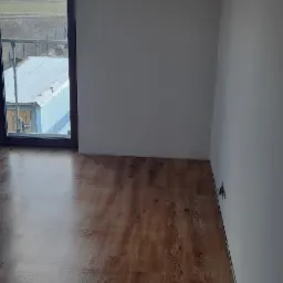 Wnętrze pokoju z panelami podłogowymi w odcieniach brązu i świeżo pomalowanymi białymi ścianami, widok przez okno na zewnątrz z metalową barierką i budynkiem gospodarczym.