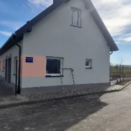 Nowo otynkowany budynek usługowy z szarą elewacją, z widocznym znakiem 'Budynek Obsługi' i ramą okienną opartą o ścianę przed montażem.