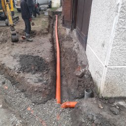 Paweł Lisowicz Usługi Ogólnobudowlane - Pomarańczowa rura kanalizacyjna ułożona w wykopie przy ścianie budynku z szarą elewacją. W tle widoczny sprzęt budowlany i osoby.