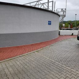 Paweł Lisowicz Usługi Ogólnobudowlane - Otynkowana, cylindryczna konstrukcja z szarą, dolną częścią i białą górną. Czerwona kostka brukowa u podstawy. Metalowe schody i platforma na górze. Pochmurne niebo.