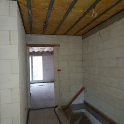 Paweł Lisowicz Usługi Ogólnobudowlane - Wnętrze w trakcie prac: ocieplone poddasze z widoczną wełną mineralną i metalową konstrukcją, ściany z białych bloczków, schody w surowym stanie i okno w tle.