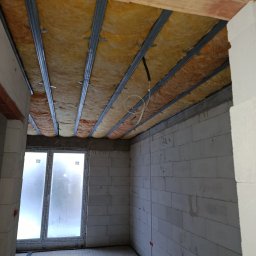 Paweł Lisowicz Usługi Ogólnobudowlane - Ocieplenie poddasza wełną mineralną, widoczne metalowe profile i instalacja elektryczna. Ściany z białego betonu komórkowego, okno w tle.
