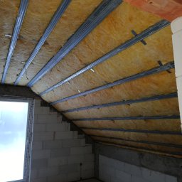 Paweł Lisowicz Usługi Ogólnobudowlane - Skośne poddasze z oknem, ocieplone wełną mineralną między metalowymi profilami. Widoczne bloczki betonowe i fragment muru z cegły. Etap prac wykończeniowych.
