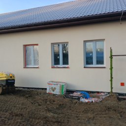 Paweł Lisowicz Usługi Ogólnobudowlane - Jasna elewacja domu jednorodzinnego z oknami zabezpieczonymi folią. Na pierwszym planie ubijarka gruntu Wacker i materiały budowlane. Widoczne rusztowanie.