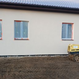 Paweł Lisowicz Usługi Ogólnobudowlane - Ściana budynku po tynkowaniu, okna zabezpieczone folią, obok stoi walec do ubijania ziemi. Widoczny fragment dachu z brązową podbitką.