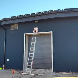 Paweł Lisowicz Usługi Ogólnobudowlane - Ciemnoszara elewacja budynku z drabiną opartą o bramę garażową. Widoczny samochód dostawczy firmy i narzędzia. Słoneczny dzień.