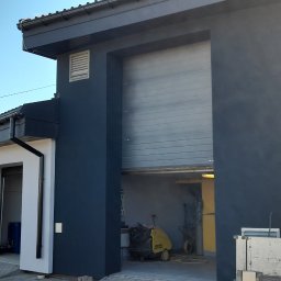 Paweł Lisowicz Usługi Ogólnobudowlane - Budynek po renowacji z otwartymi bramami garażowymi, pomalowany na szaro, z widocznym samochodem w tle i sprzętem wewnątrz garażu.