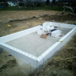 Fundament budynku w trakcie budowy, obłożony białymi panelami izolacyjnymi z tworzywa sztucznego, widoczne narzędzia i materiały budowlane wewnątrz fundamentu, w tle krajobraz wiejski z domami...