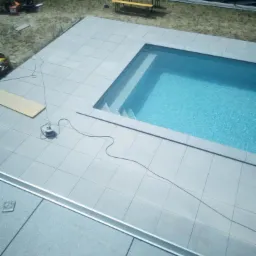 Widok z góry na nowo wyłożony, jasnoszarymi płytkami taras wokół basenu z błękitną wodą, z widocznym rozłożonym kablem elektrycznym i elementami wyposażenia placu budowy w tle.