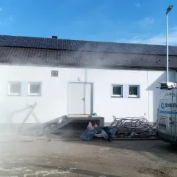 Biały budynek z panelami słonecznymi na dachu w trakcie czyszczenia elewacji pod ciśnieniem, widoczny samochód serwisowy i rusztowania.