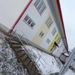 Budynek mieszkalny po remoncie elewacji, z białymi ścianami, czerwonymi rynnami i fragmentem żółtego pasa, widoczny z poziomu schodów pokrytych śniegiem.