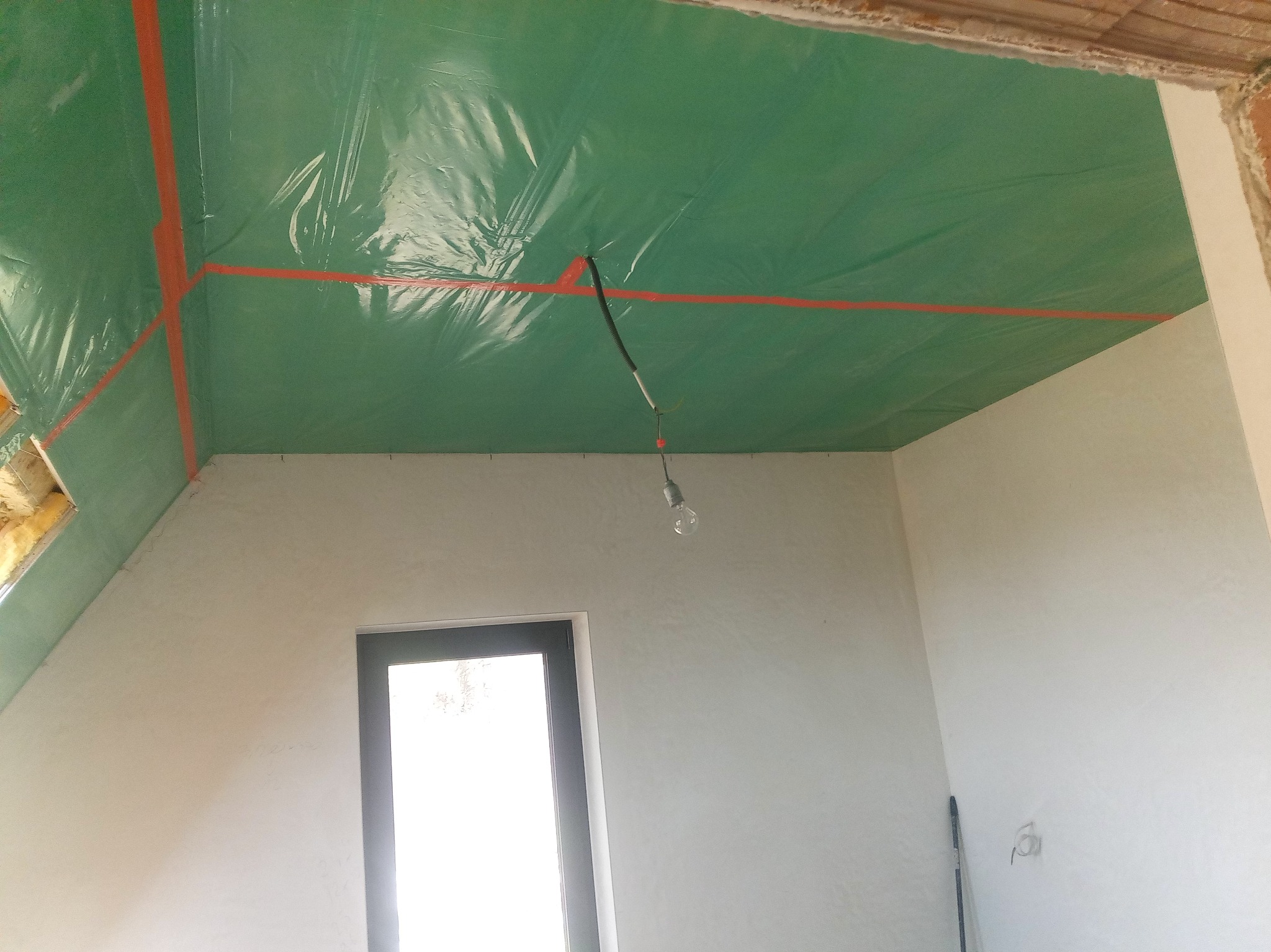 Poddasze w trakcie prac: zielona folia paroizolacyjna zabezpieczona czerwoną taśmą na suficie, surowa ściana z oknem i wisząca żarówka.