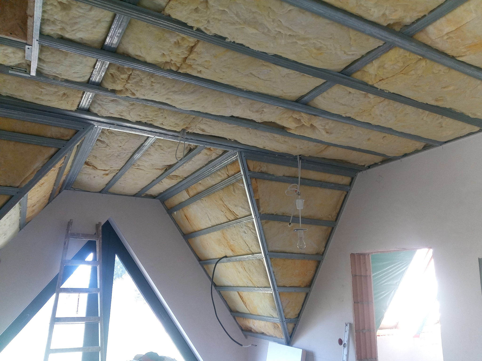 Poddasze w trakcie ocieplania wełną mineralną, widoczna metalowa konstrukcja sufitu podwieszanego, fragment okna dachowego i niedokończone ściany.