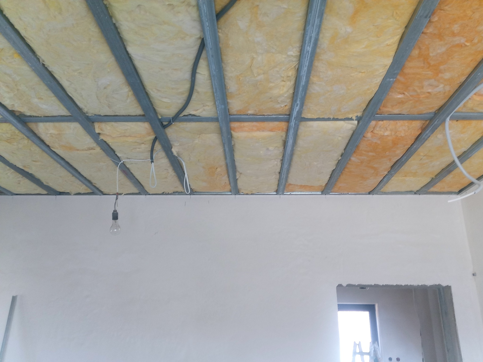 Sufit podwieszany w trakcie montażu: widoczna metalowa konstrukcja wypełniona wełną mineralną, wisząca żarówka, surowe ściany z otworem drzwiowym w tle.