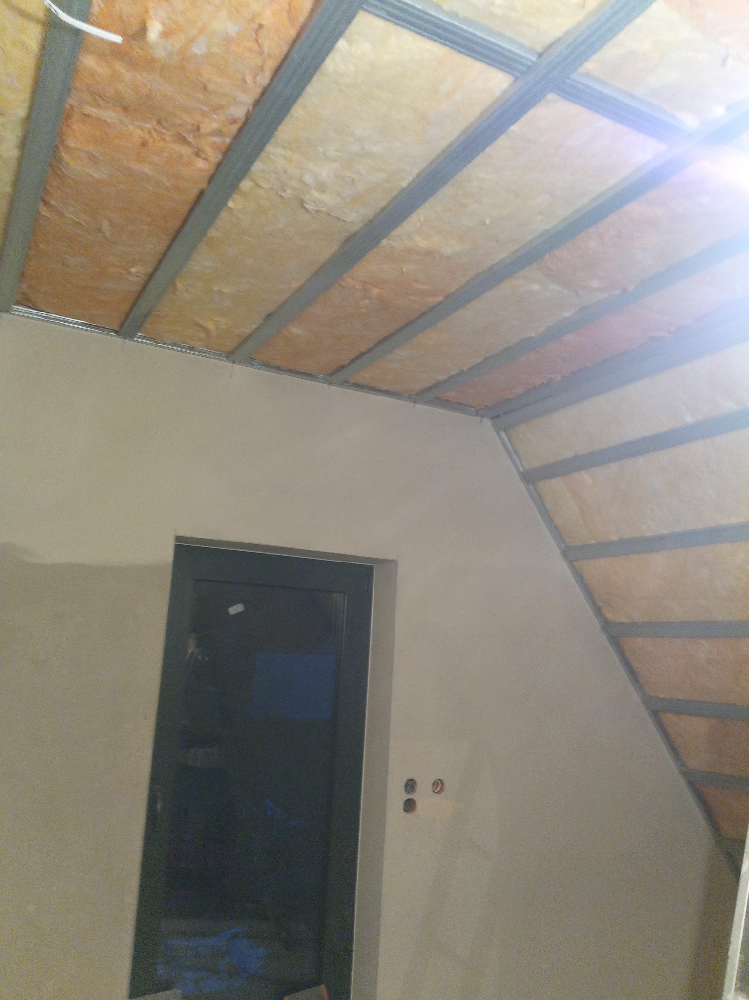 Skośny sufit poddasza z widoczną izolacją z wełny mineralnej pomiędzy metalowymi profilami, ściana ze świeżym tynkiem, widoczne puszki elektryczne i ciemne drzwi.