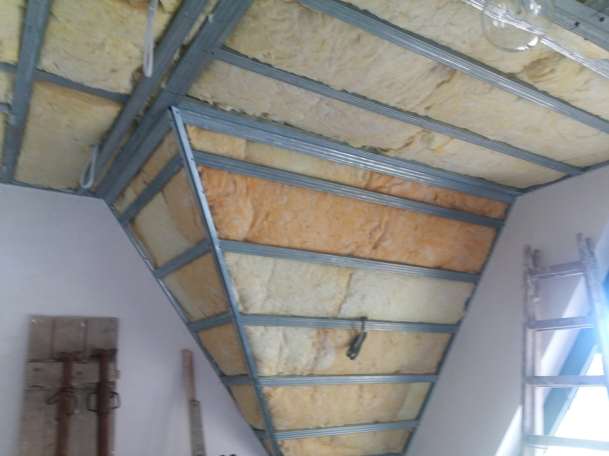 Skośne poddasze w trakcie ocieplania, widoczna wełna mineralna pomiędzy metalowymi profilami, drabina oparta o ścianę, narzędzia budowlane oparte o ścianę.