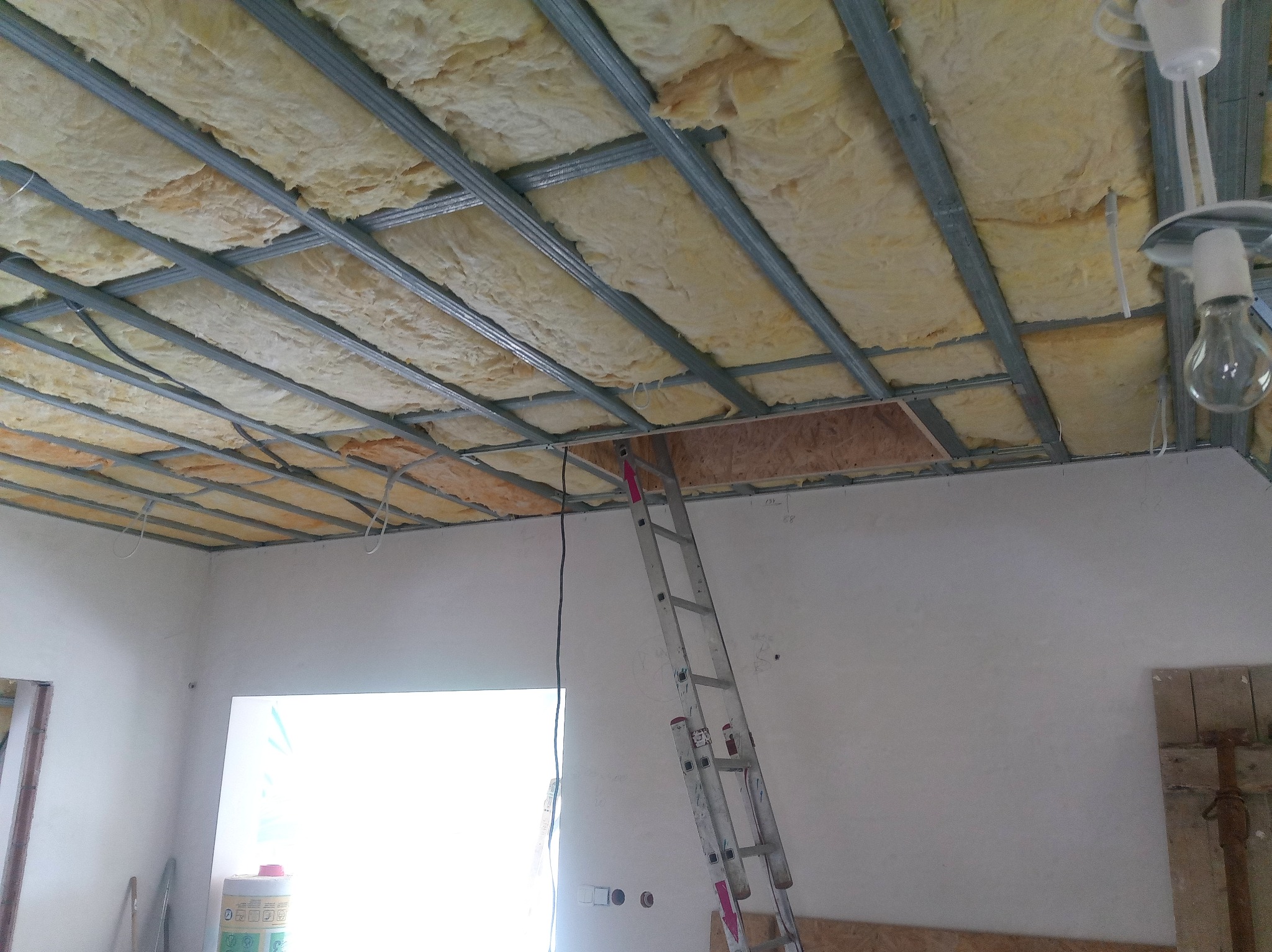 Montaż izolacji z wełny mineralnej w metalowym stelażu sufitu podwieszanego, widoczna drabina oparta o ścianę i prowizoryczne oświetlenie z żarówką.