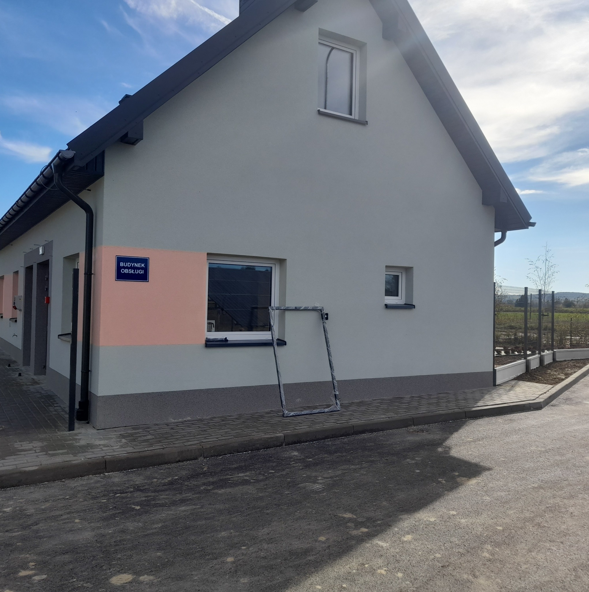 Nowo otynkowany budynek usługowy z szarą elewacją, z widocznym znakiem 'Budynek Obsługi' i ramą okienną opartą o ścianę przed montażem.