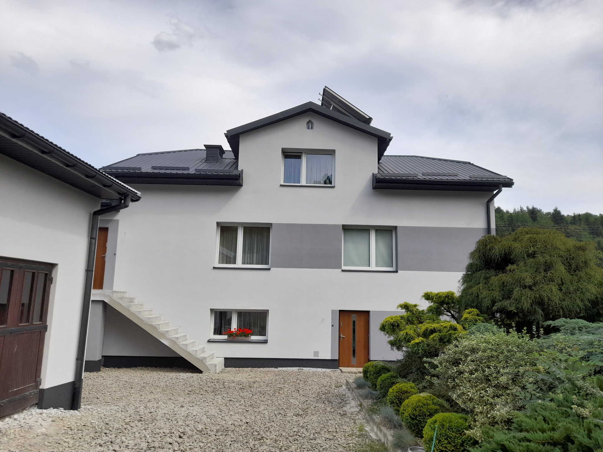 Dwukondygnacyjny dom z szarą elewacją, ciemnym dachem i schodami zewnętrznymi. Widoczne okna z kwiatami, zadbana zieleń i żwirowe podłoże. Ujęcie z poziomu gruntu.