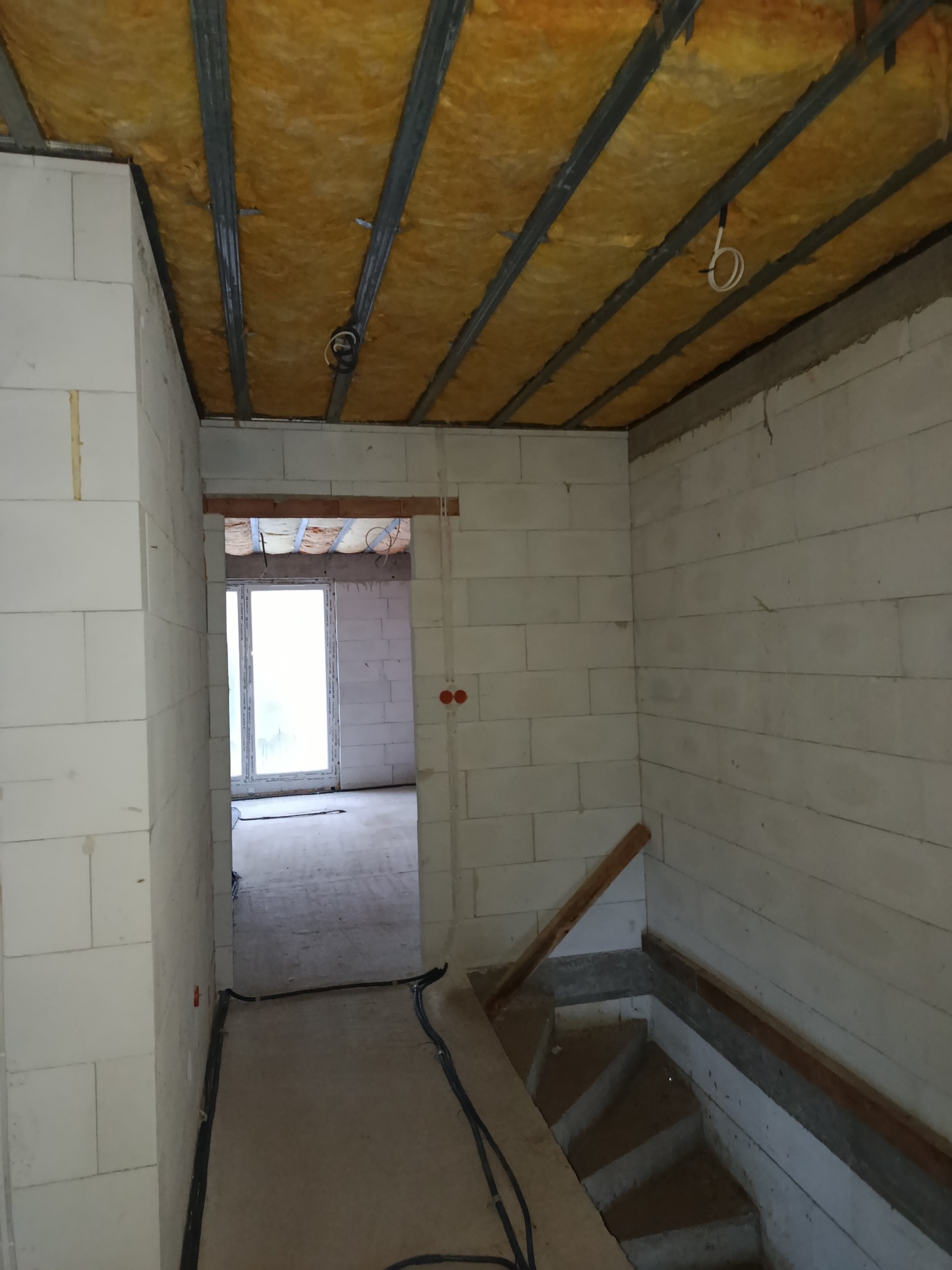 Wnętrze w trakcie prac: ocieplone poddasze z widoczną wełną mineralną i metalową konstrukcją, ściany z białych bloczków, schody w surowym stanie i okno w tle.
