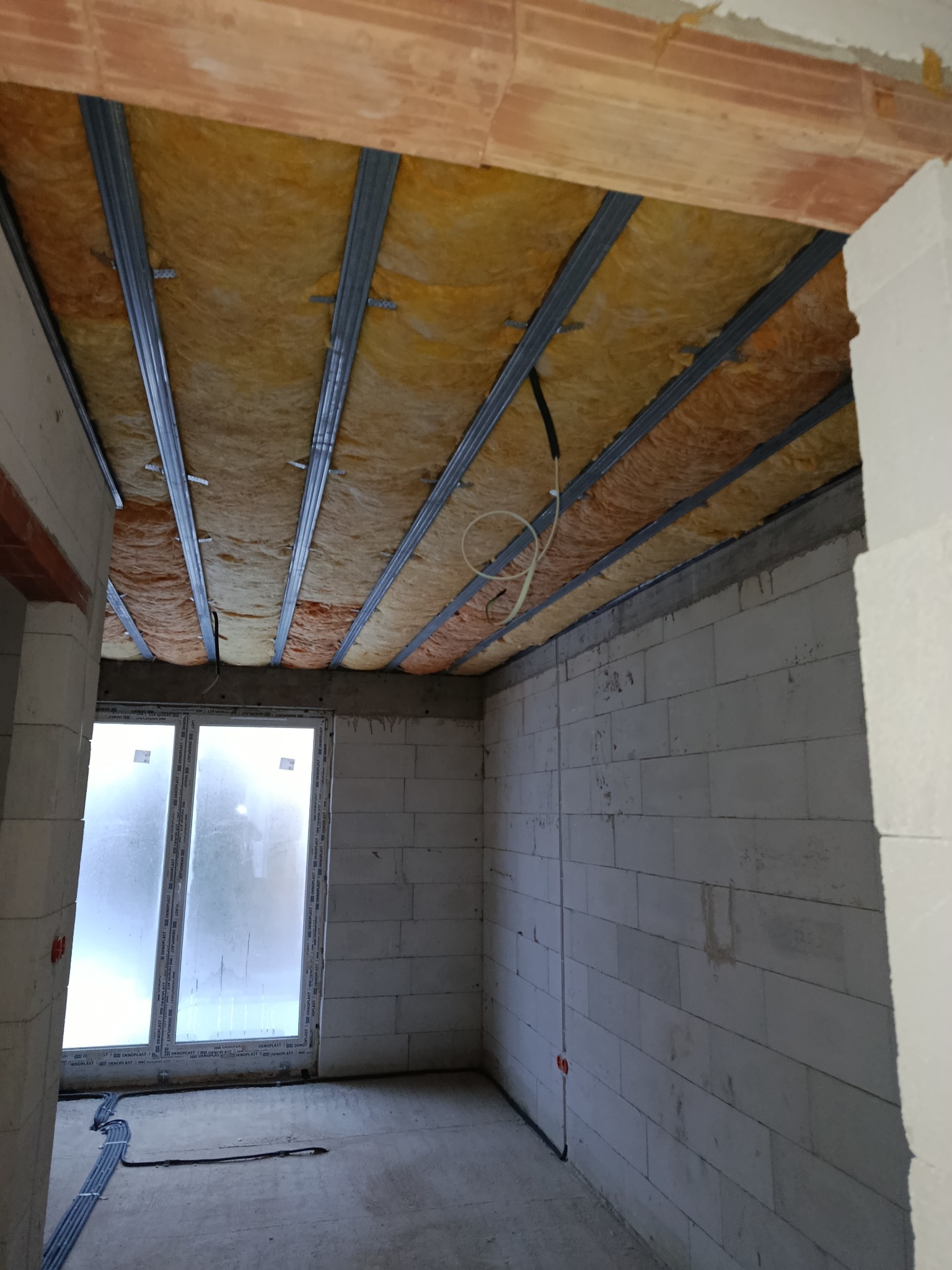 Ocieplenie poddasza wełną mineralną, widoczne metalowe profile i instalacja elektryczna. Ściany z białego betonu komórkowego, okno w tle.