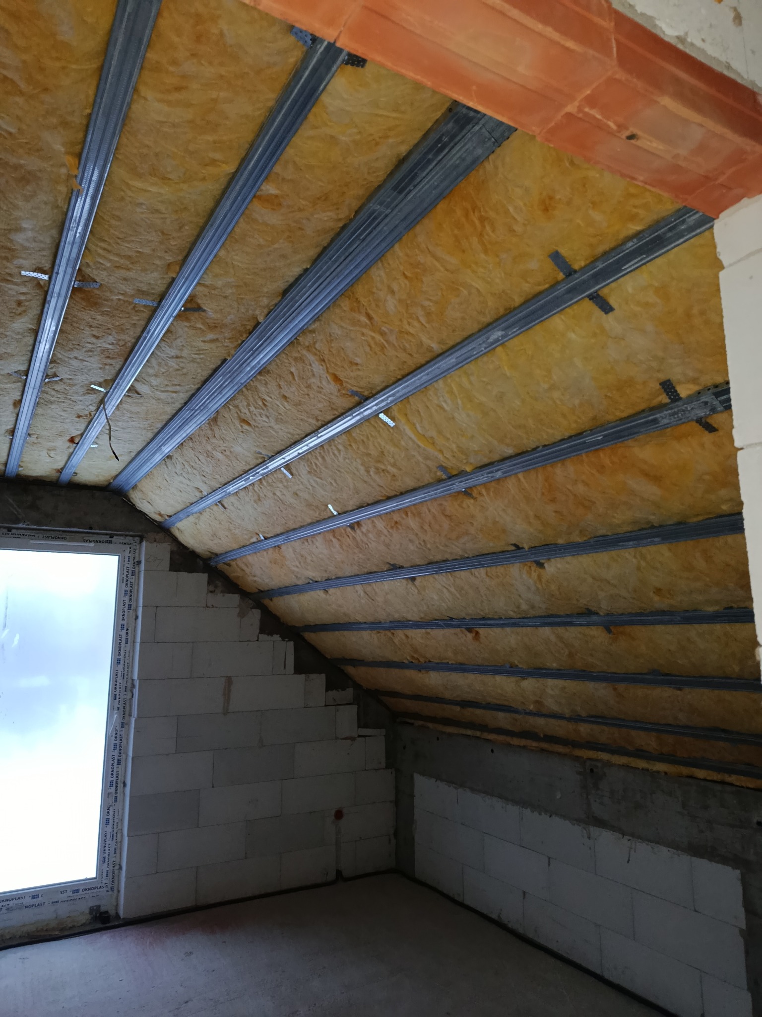Skośne poddasze z oknem, ocieplone wełną mineralną między metalowymi profilami. Widoczne bloczki betonowe i fragment muru z cegły. Etap prac wykończeniowych.